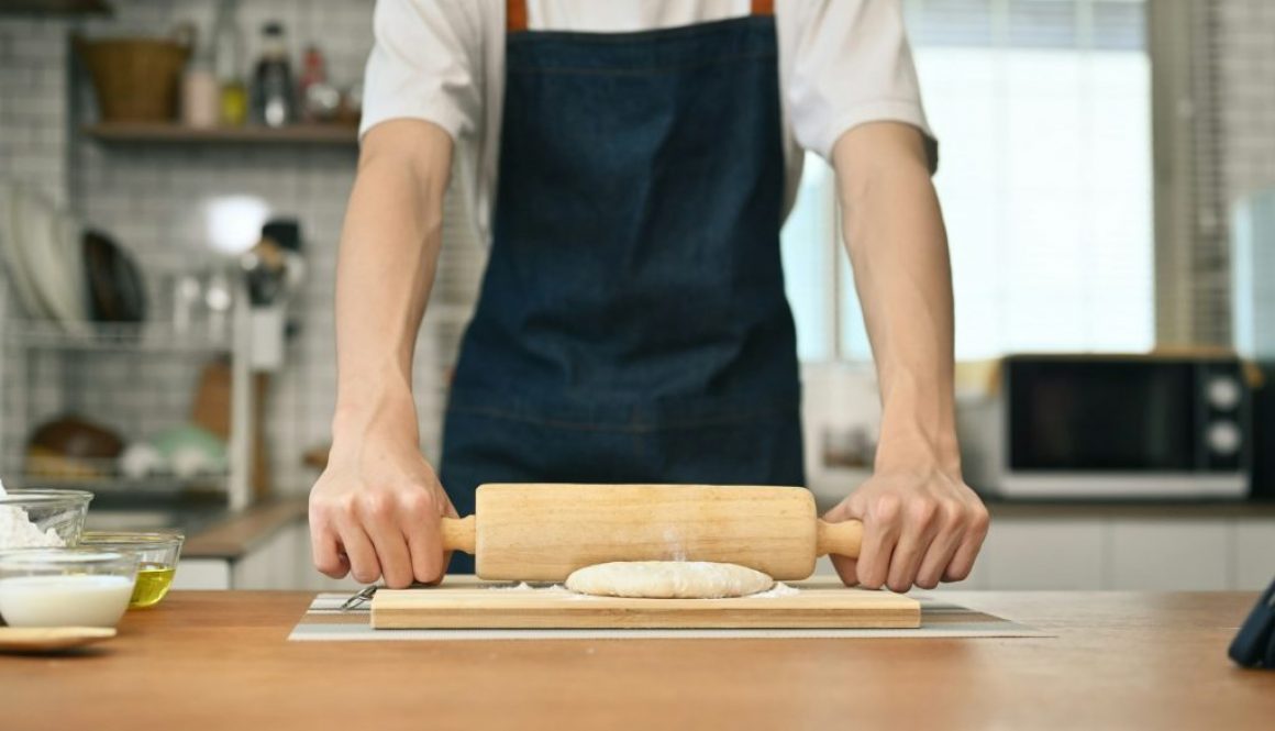 Man wearing aprons preparing homemade pastry, kneading dough with a rolling pin on wooden table.